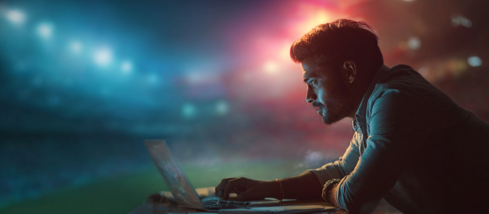 Focused man using his laptop for Ywkk sports betting with a glowing stadium in the background.